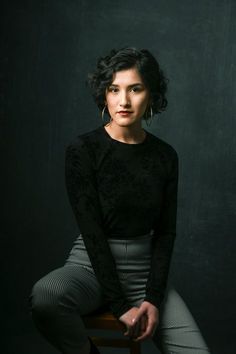 a woman sitting on top of a wooden chair in front of a black wall with her legs crossed