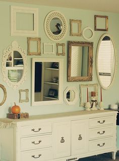 a white dresser topped with lots of mirrors next to a wall filled with framed pictures