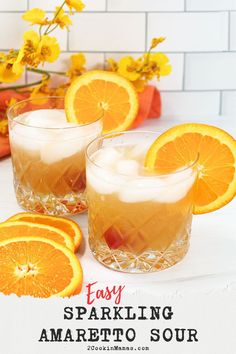 two glasses filled with oranges and ice sitting on top of a table next to flowers