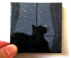 a hand holding up a small square painting with a black cat in the window at night