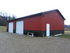 a red and black building with two garages next to it on gravel road in front of trees