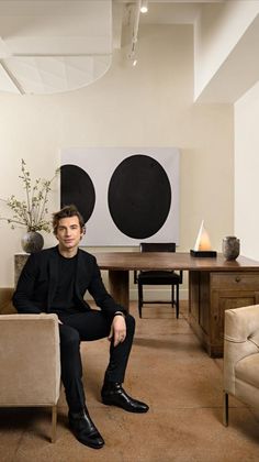 a man sitting in a living room next to a table and chair with a vase on it