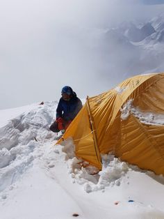 a man sitting next to a tent on top of a snow covered mountain with mountains in the background