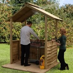 a man and woman standing in the grass near a small wooden structure with a grill on it