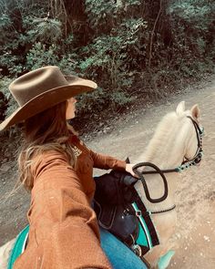 a woman riding on the back of a white horse down a dirt road with trees in the background