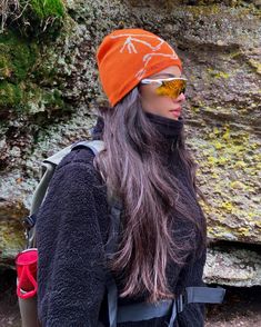 a woman with long hair wearing a hat and sunglasses standing in front of a rock wall