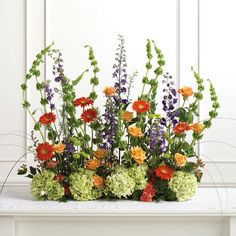 an arrangement of flowers on a table in front of a white wall with green and orange accents