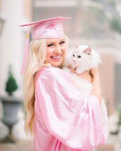 a woman in a pink graduation gown holding a white cat and smiling at the camera