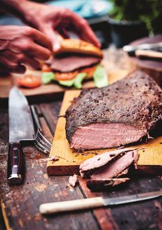 a person cutting meat on top of a wooden cutting board next to knifes and forks