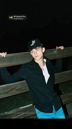 a young man leaning on a wooden fence with his hands in the air while wearing a baseball cap