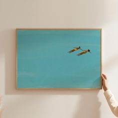 two birds flying in the sky above a person's hand holding up a painting