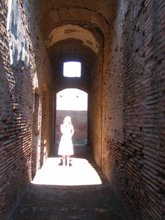 a woman standing in an alley between two brick buildings with sunlight coming through the windows