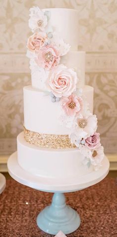 a three tiered white cake with pink flowers on top and gold glitters around the edges