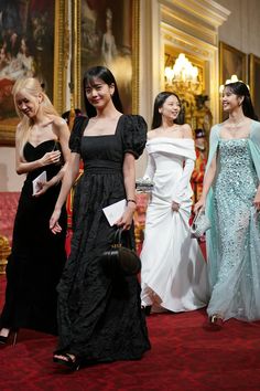 four women in evening gowns are walking down the red carpeted hall, one is holding a purse