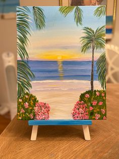 an easel painted with palm trees and pink flowers on the beach at sunset or sunrise