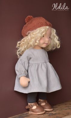 a doll with blonde hair wearing a gray dress and brown hat is standing on a shelf