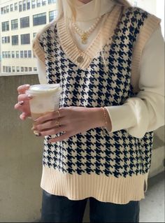 a woman wearing a sweater vest and holding a cup in her hand while standing next to a tall building