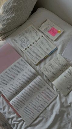 several books are laying on a bed with white sheets and pillows, next to an open notebook