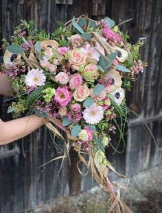 a bridal bouquet with pink flowers and greenery in front of a wooden fence