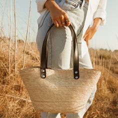 Easily pack up the Jude tote in a suitcase so you can enjoy its versatility with the outfits you've packed for your travels. Its unique shape, with neutral design elements make it the bag that goes with everything. Each handmade basket is unique and may have small imperfections or vary in size and color. All Leah products are responsibly sourced and ethically crafted. Handles: Double layer black leather handles No interior lining Length: 9" Width: 16" Height: 8" Handle Drop: 10" Imported Beige Rectangular Straw Bag For Travel, Casual Beige Basket Bag, Casual Weekender Tote Bag, Trendy Beige Beach Bag For Travel, Casual Beige Straw Bag For Travel, Casual Beige Straw Travel Bag, Trendy Beige Weekender Bag For Travel, Casual Weekender Tote Bag With Top Handle, Chic Natural Satchel Bucket Bag