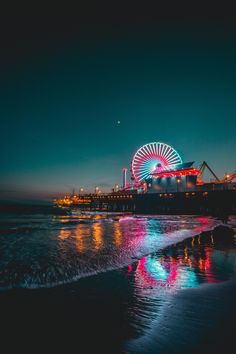 the ferris wheel is lit up at night on the beach by the water's edge