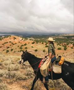 a man riding on the back of a black horse through a desert landscape with clouds in the background