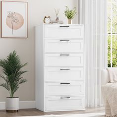 a white chest of drawers next to a plant in a living room with a window