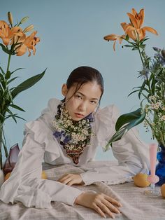 a woman sitting at a table with flowers in front of her and an arrangement of vases behind her