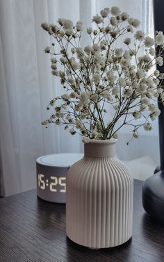 a white vase with flowers in it sitting on a wooden table next to a clock