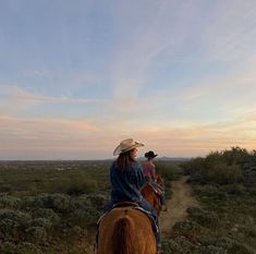 two people riding horses on a dirt path in the middle of an open field at sunset