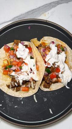 two tacos with meat, sour cream and vegetables on a black plate sitting on a marble counter top