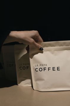 a person's hand holding a coffee bag on top of a table with two bags in front of them