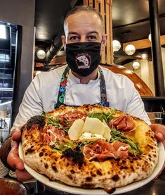 a man wearing a face mask holding a large pizza on a white plate with toppings