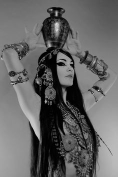 a woman with long black hair holding a vase on her head and wearing bracelets