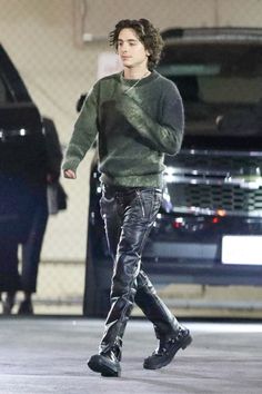 the young man is walking down the street wearing black pants and a green sweater with his hands in his pockets