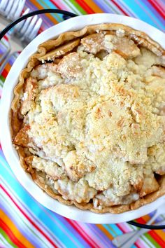 an apple pie is in a white bowl on a colorful tablecloth with utensils