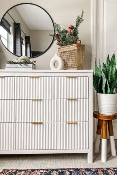 a white dresser sitting next to a mirror and potted plant