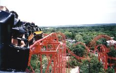 people are riding the roller coaster at an amusement park with trees and buildings in the background