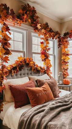 a bed covered in lots of pillows next to two windows with fall leaves on them