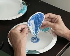 a person is painting a plate with blue and white designs on the rim, while another hand holds an object in front of them