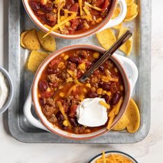 two bowls of chili with cheese and tortilla chips on a tray next to a bowl of sour cream