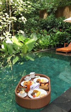 a wicker boat filled with food sitting on top of a pool surrounded by greenery