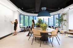 two women are writing on the whiteboard in an office with large windows and potted plants