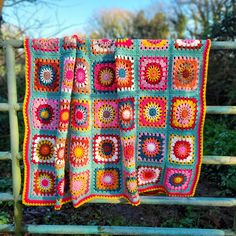 a crocheted blanket is hanging on a wooden rail in front of some bushes