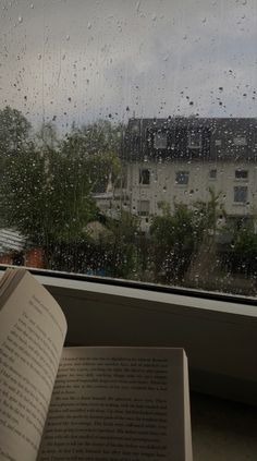 an open book sitting on top of a window sill next to a rain covered window