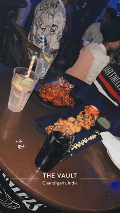 a table topped with plates of food next to a drink and two people in the background