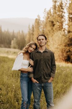 a man and woman are standing in the grass near some trees, one is holding his arm around the other's shoulder