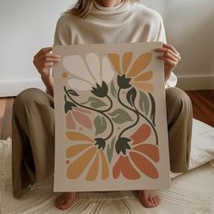 a woman sitting on the floor holding up a poster with an image of flowers painted on it