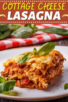 a white plate topped with lasagna covered in cheese and basil leaves next to a red and white checkered table cloth