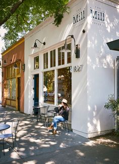 a woman sitting on a chair in front of a building with tables and chairs outside